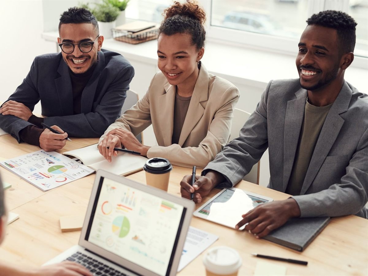 Três profissionais sorrindo durante uma reunião, com notebook e relatórios à frente